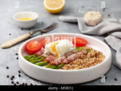 Una sana prima colazione. Asparagi al forno con pancetta, quinoa, uovo cotto in una ciotola bianco su una pietra grigia sullo sfondo Foto Stock