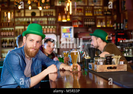 Vita-up verticale di bello uomo barbuto che indossa jeans e maglietta verde Bowler cappello in posa per la fotografia mentre è seduto al banco bar e celebrando Foto Stock