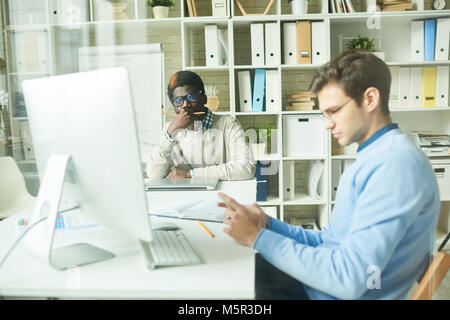 Processo di lavoro in modern open plan office: multi-etnico team di lavoratori di colletto bianchi seduti alla scrivania e lavorando su start-up progetti, vista attraverso Foto Stock