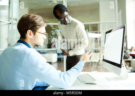 Giovani manager in occhiali seduto davanti al computer puntando alla schermata vuota mentre mostrando qualcosa al suo americano africano collega, interi Foto Stock