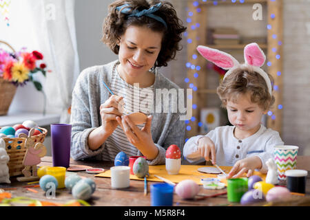 Ritratto di carino piccolo ragazzo indossa orecchie di coniglietto di pittura delle uova di Pasqua in casa con la mamma mentre facendo i preparativi per le vacanze Foto Stock