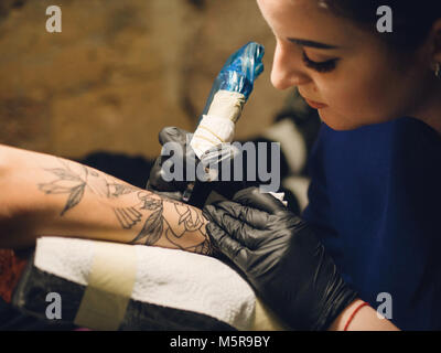 Ritratto di bruna tattooer femmina in viola abbigliamento rendendo tattoo sull'uomo del braccio del suo client con la pistola in studio. Giovane donna di affari in opere Foto Stock