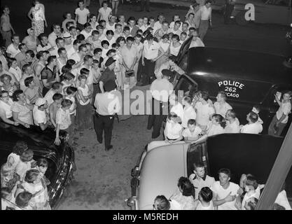 Una folla di vicinato orologi come il corpo della vittima di omicidio viene caricato nel retro della polizia funebre nel lato sud di Chicago, ca. 1955. Foto Stock