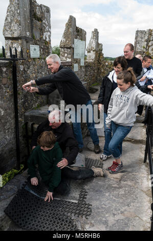 Un giovane visitatore guardato dalla sua famiglia mentre si distende a baciare la pietra Blarney con l'aiuto di un membro del personale in cima al castello di Blarney Foto Stock