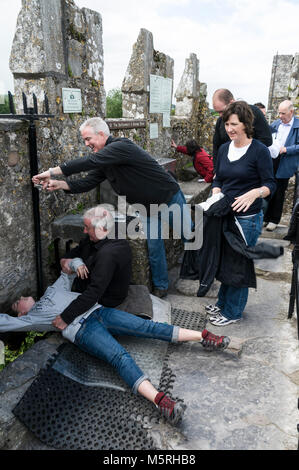 Un giovane visitatore guardato dalla sua famiglia mentre si distende di nuovo a baciare la pietra Blarney con l'aiuto di un membro del personale in cima al castello di Blarney Foto Stock