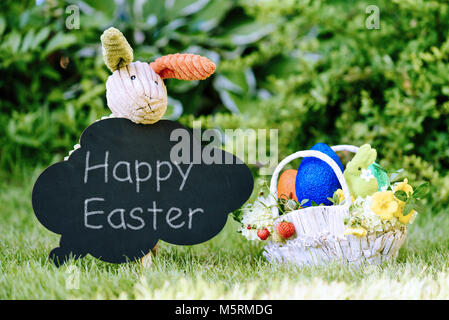 Toy bunny con chalk testo 'Felice Pasqua' sulla lavagna e cesto con uova, fiori e frutti di bosco Foto Stock