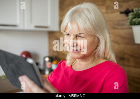 Ritratto di felice senior lady in abito rosso utilizzando tablet in cucina Foto Stock