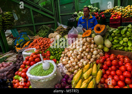 Un fornitore colombiano, tenendo un mais, sta dietro le pile di verdure al mercato di Paloquemao di Bogotá, in Colombia. Foto Stock