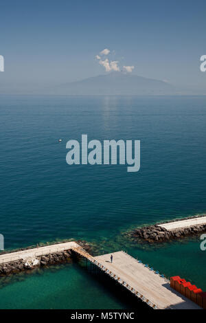 Vista del Monte Vesuvio guardando attraverso la baia di Napoli da Sorrento con una singola persona su un molo con la spiaggia rossa di capanne. Foto Stock