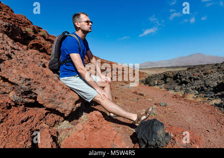 Un uomo con uno zaino seduto su di una roccia rossa e il riposo durante il trekking nel paesaggio vulcanico del Parco Nazionale Timanfaya su Lanzarote, il canarino Islan Foto Stock