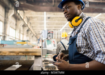 Concentrato African American Lavoratore che indossa in generale e di compressori hardhat macchina operatrice con l aiuto della tavoletta digitale, interno della spaziosa partono di produzione Foto Stock
