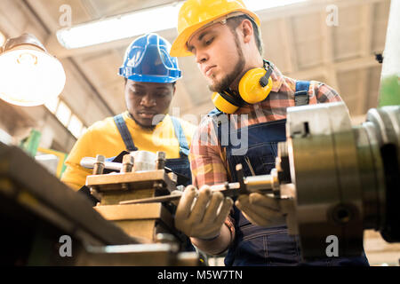 Basso angolo vista del tornio concentrato operatore che indossa in generale e di compressori hardhat distratti dal lavoro al fine di rispondere alle domande di afro-americano di ispezione Foto Stock