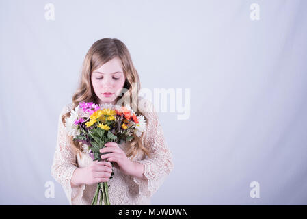 Twelve-year-old girl with long, dirty blonde hair looking down at colorful bouquet of daisies against white background Foto Stock
