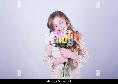 Sorridente bambina di dodici anni con lunghi, sporchi capelli biondi abbracciando colorato bouquet di margherite con gli occhi chiusi contro uno sfondo bianco Foto Stock