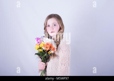 12-anno-vecchia ragazza con lunghi, sporchi capelli biondi ruotata lateralmente holding colorato bouquet di margherite, occhi aperti, triste espressione, sfondo bianco Foto Stock