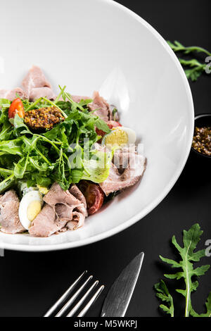 Insalata con un arrosto di manzo, rucola, pomodori ciliegini, uova e grano di senape su uno sfondo nero. close up Foto Stock