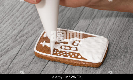 Close-up di femmine della mano mentre la decorazione di carino gingerbread su tavola di legno. Pasticceria la pittura è una chiesa sul cookie utilizzando sacchetto di glassa con la glassa. Foto Stock