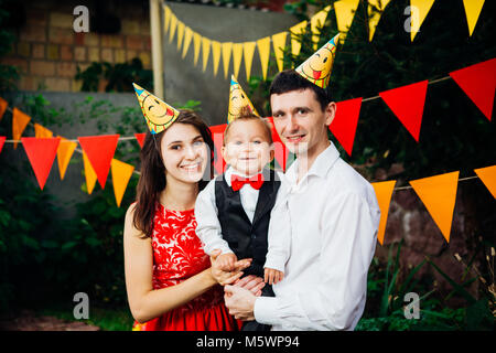 Tema bambini festa di compleanno. Padre di famiglia e madre figlio di contenimento di un anno sullo sfondo di verde e di decorazioni festive, ghirlande e colorato Foto Stock