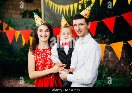 Tema bambini festa di compleanno. Padre di famiglia e madre figlio di contenimento di un anno sullo sfondo di verde e di decorazioni festive, ghirlande e colorato Foto Stock