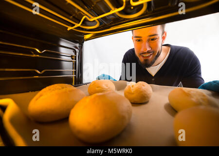 Felice giovane uomo che indossa guanti blu estraendo il vassoio di biscotti dal forno Foto Stock