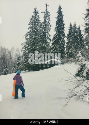 Una ragazza di snowboard in un casco blu sorge con la sua mano sul lato di un arancio brillante pensione nella foresta e guarda a un vecchio abbandonato casa in legno h Foto Stock