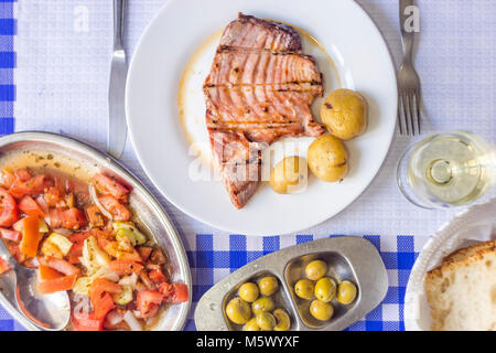 Bistecca di tonno accompagnata con patate, olive, insalata di pomodoro, pane e vino sulla piastra bianca Foto Stock