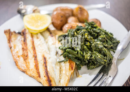 Grigliata di branzino servito con limone, patate al forno e spinaci su piastra bianca Foto Stock