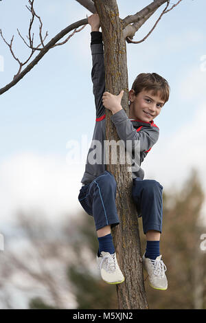 Il 10-anno-vecchio ragazzo arrampicata su un albero che guarda verso la telecamera. Inquadratura orizzontale con luce naturale Foto Stock