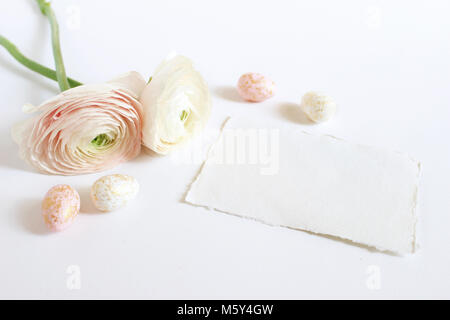 Molla della scheda Messaggi di saluto, invito con rosa e bianco golden spotted uova pasquali roselline, ranuncoli fiori e vuoto di carta craft giacente sul tavolo bianco. Stile femminile stock photo. Foto Stock