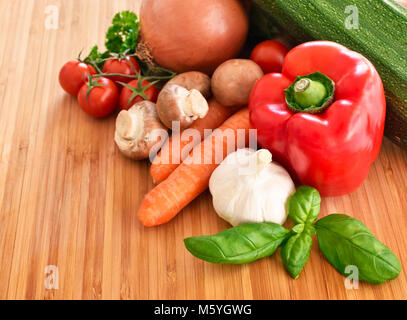 Fresche verdure crude su un tagliere. disposizione di peperone rosso, zucchine, broccoli, insalata, cipolla, carote e aglio, decorate in bambù. Foto Stock