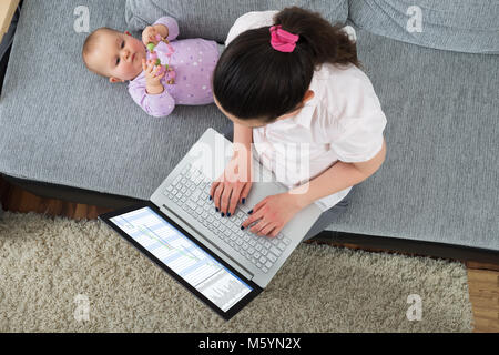 Donna seduta su un divano con computer portatile mentre il suo bambino gioca con Toy Foto Stock