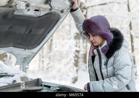 La ragazza ha sollevato il cofano dalla vettura e guarda al motore. Inverno, giorno. Foto Stock