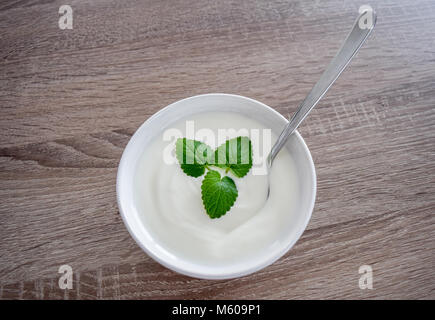 Vaso in ceramica di yogurt bianco con foglie di menta come decorazione e cucchiaio all'interno isolato su vintage tavola in legno rustico da vista superiore, yogurt naturale. yog Foto Stock