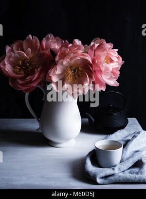 Il fascino di corallo le peonie in piena fioritura, in una caraffa del bianco sul tavolo con tazza e teiera tetsublin, sfondo scuro, colpo di luce naturale Foto Stock