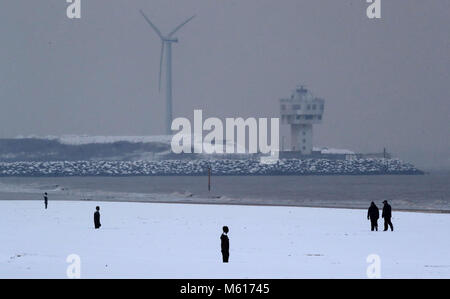 Walkers nella neve vicino a un altro luogo da Anthony Gormley, su Crosby il faggio vicino al Liverpool, a seguito di una notte pesante nevicata che ha provocato interruzioni in tutta la Gran Bretagna. Foto Stock