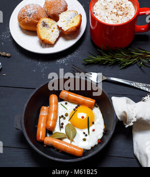 Uova fritte con salsicce in un nero padella, cacao con marshmallows e muffin su tavoli in legno nero, vista dall'alto Foto Stock