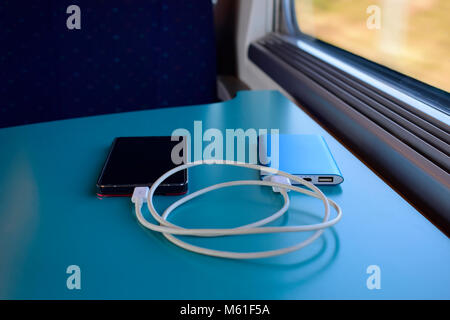 Nero smartphone carica con il blu della banca di alimentazione blu su sfondo tabella vicino la sede all'interno del treno contro il sole attraverso la finestra. Foto Stock