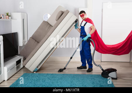 Bidello maschio indossando il costume del supereroe di pulizia pavimento in legno con aspirapolvere in soggiorno Foto Stock