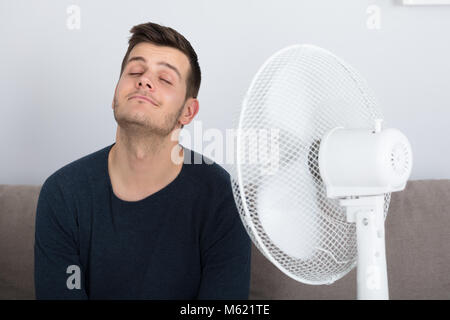 Giovane uomo seduto sul lettino godendo brezza con ventola elettrica a casa Foto Stock