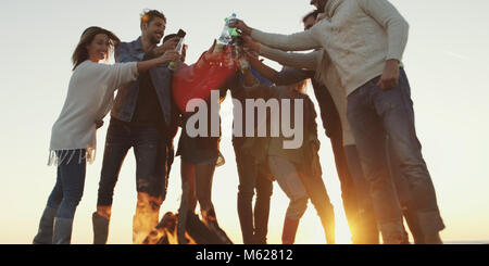 Giovani amici fare un brindisi con la birra e divertirsi intorno al falò presso il beach party sulla giornata autunnale Foto Stock