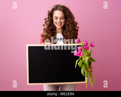 Rosa umore. sorridente giovane donna con lunghi mossi brunette capelli su sfondo rosa con un bouquet di tulipani guardando a bordo nero Foto Stock