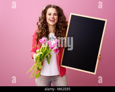 Rosa umore. sorridente giovane donna con lunghi mossi brunette capelli isolati su rosa con un bouquet di tulipani che mostra la scheda di colore nero Foto Stock