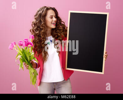 Rosa umore. sorridente donna elegante con lunga ondulato brunette capelli su sfondo rosa con un bouquet di tulipani guardando a bordo nero Foto Stock