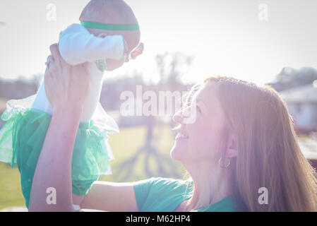 Primo piano di una madre sorridente sollevando il neonato in condizioni di luce solare intensa per la festa di San Patrizio Foto Stock