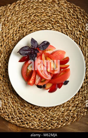 Insalata di pomodoro e prugne rosse con basilico per una dieta sana. Low-calorie cibo per il controllo del peso corporeo. Snack veloce. Su un tavolo di legno, un cavalletto di vimini. Foto Stock