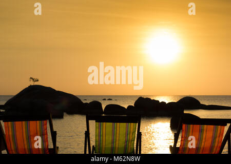 Vuoto tre sedie a sdraio di fronte al mare al tramonto con silhouette di rocce su acqua e albero singolo sulla parte superiore di quella grande in Koh Phangan, Thailandia Foto Stock