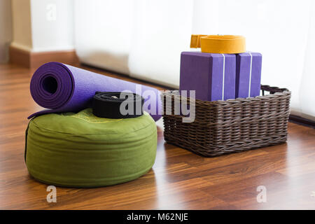 Vari i sostegni di yoga studio sul pavimento di legno. Set di blocchi in cesto in vimini, cinghie, tappetino e cuscino verde nel centro yoga. Wellness nozione di attività Foto Stock