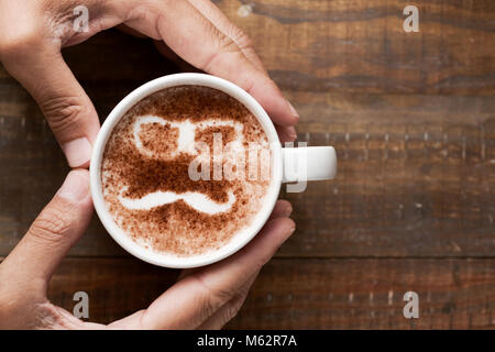 Giovane uomo formando un cuore con le mani attorno a una tazza di cappuccino, con un paio di occhiali e un baffi disegnati con il cacao in polvere sulla sua schiuma, su Foto Stock