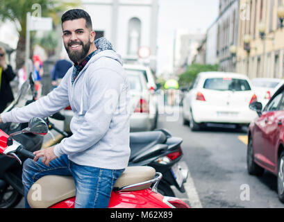 Felice giovane parcheggio ciclomotore rosso nella città vecchia di Las Palmas, Spagna. Piena barbuto hipster seduti sul classico italiano in stile retrò motocicletta sulla strada Foto Stock