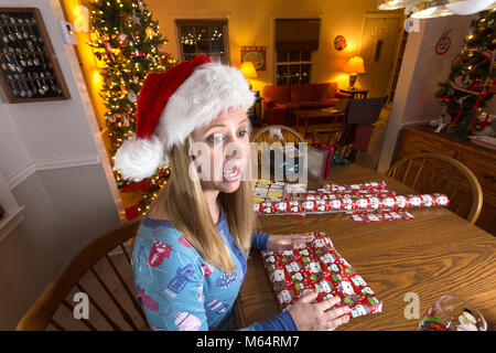 Giovane donna caucasica avvolge i regali di Natale mentre si beve il vino e indossando pigiami nel suo soggiorno Foto Stock